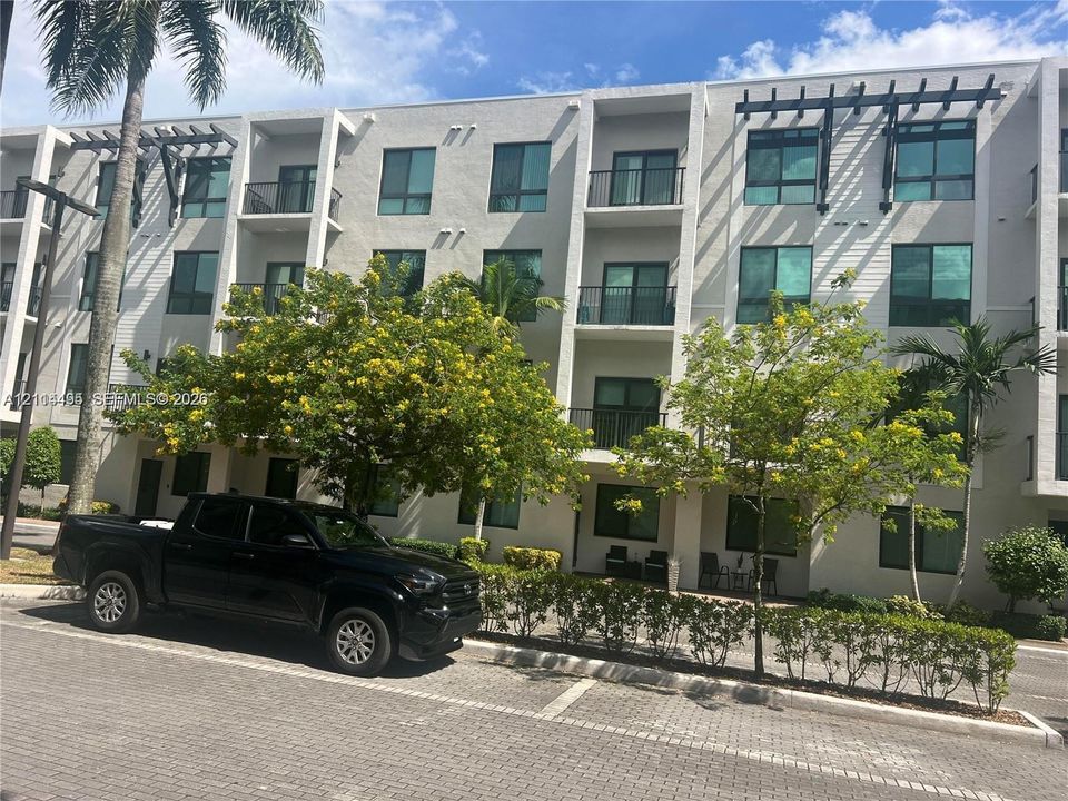 Multi-story stucco exterior featuring private balconies with decorative timber pergolas, large glass window panels, and paver-stone parking surfaces