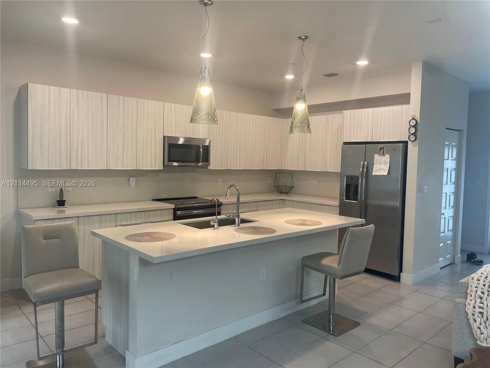 Kitchen featuring textured white cabinetry, solid surface countertops, a central island with an integrated sink, stainless steel appliances, and dual pendant lighting