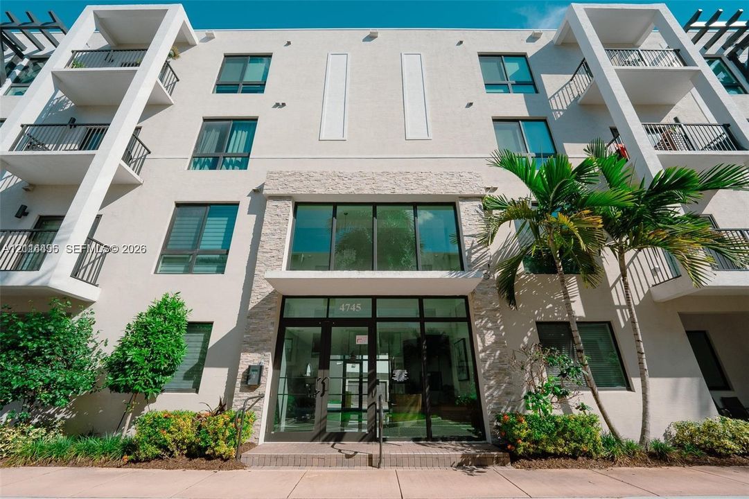Stucco exterior facade featuring multi-level balconies with black metal railings, floor-to-ceiling glass entryway, and stone-veneer cladding