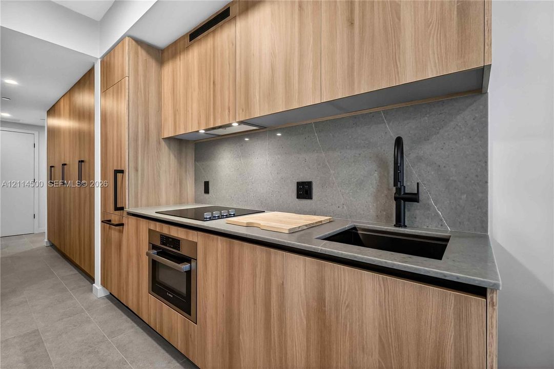 Modern kitchen area featuring flat-panel wood-finish cabinetry, stone-surface countertops, full-height stone backsplash, integrated stainless steel oven, and a black glass electric cooktop