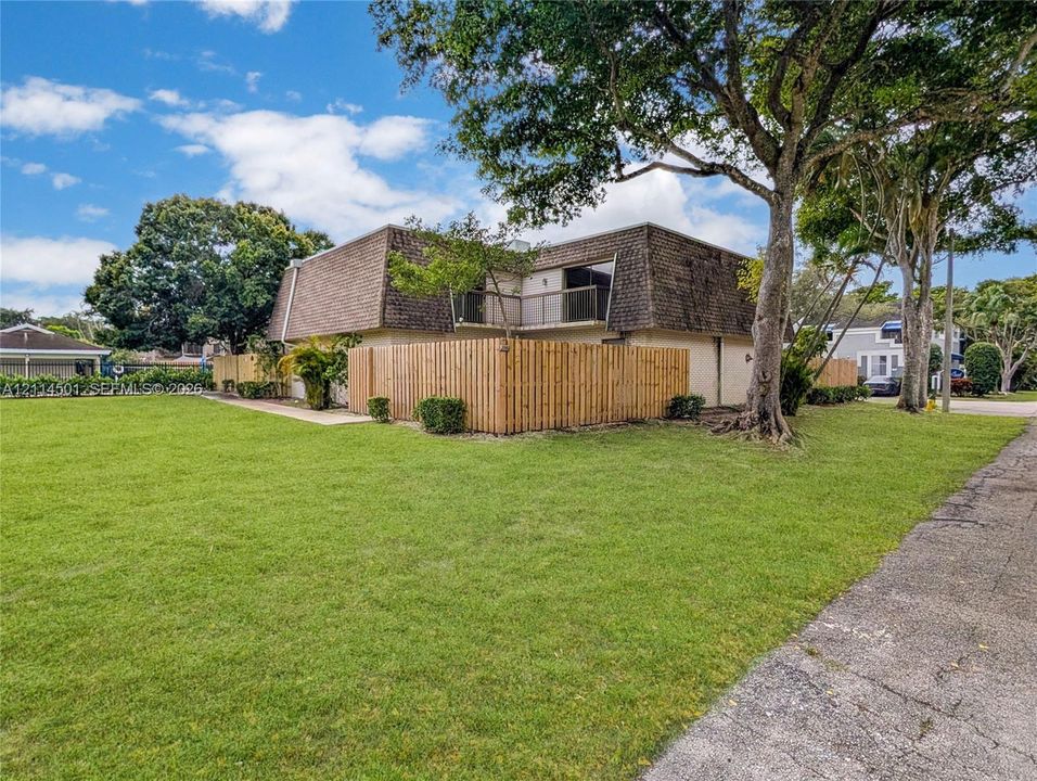 Exterior profile featuring a mansard roofline, wood-slat privacy fencing, balcony access, and mature trees on a grass lawn