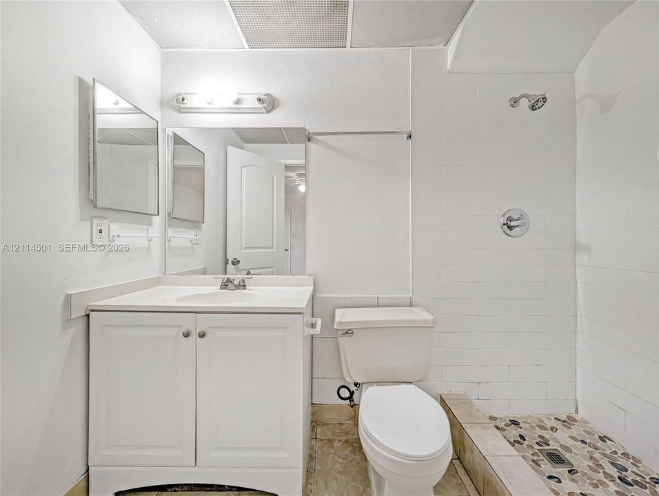 Bathroom featuring a white vanity cabinet, rectangular mirror, walk-in shower with white tile surround, and decorative pebble-style stone flooring