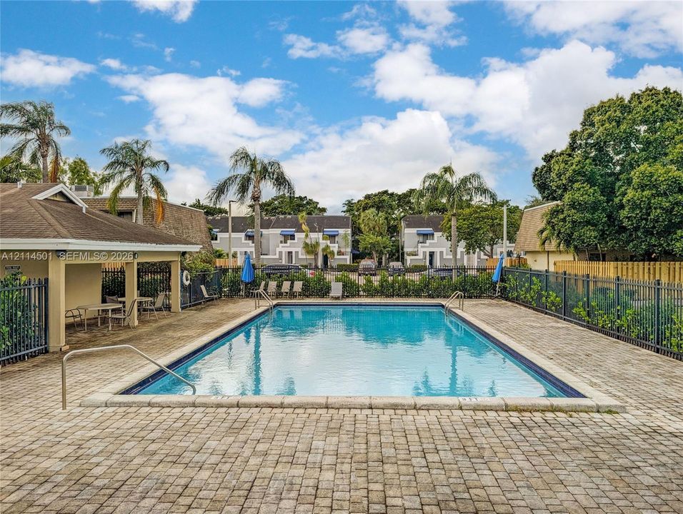 Community swimming pool featuring a rectangular basin, stone-paver pool deck, covered pavilion, and perimeter metal fencing