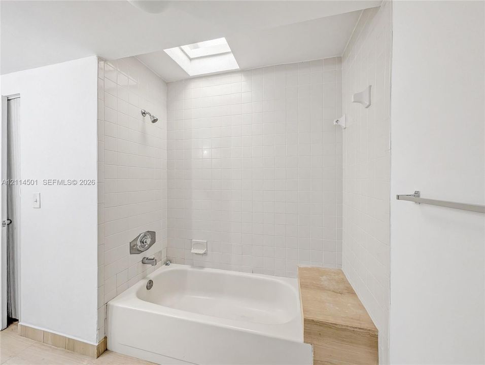 Bathtub with white tile surround, integrated stone-finish bench, and ceiling skylight