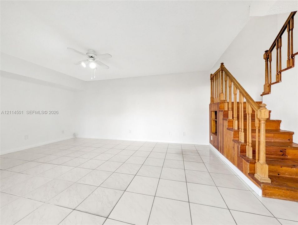 Open-concept space featuring light-toned tile flooring, a ceiling fan with integrated lighting, and a prominent wood staircase with matching balusters and built-in shelving