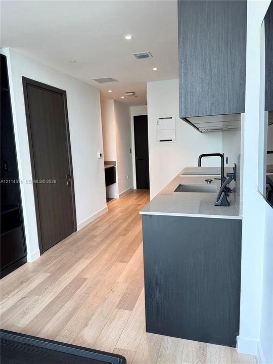 Kitchenette featuring wood-finish flooring, dark-toned cabinetry, a rectangular stainless steel sink with a matte black faucet, and a quartz countertop