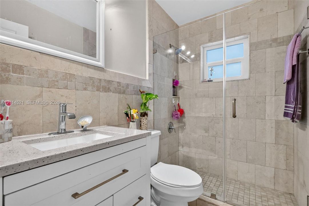 Bathroom featuring a white vanity with a light-toned stone countertop, square undermount sink, and metallic hardware, complemented by a walk-in shower with floor-to-ceiling tile, a frameless glass door, and integrated shelving