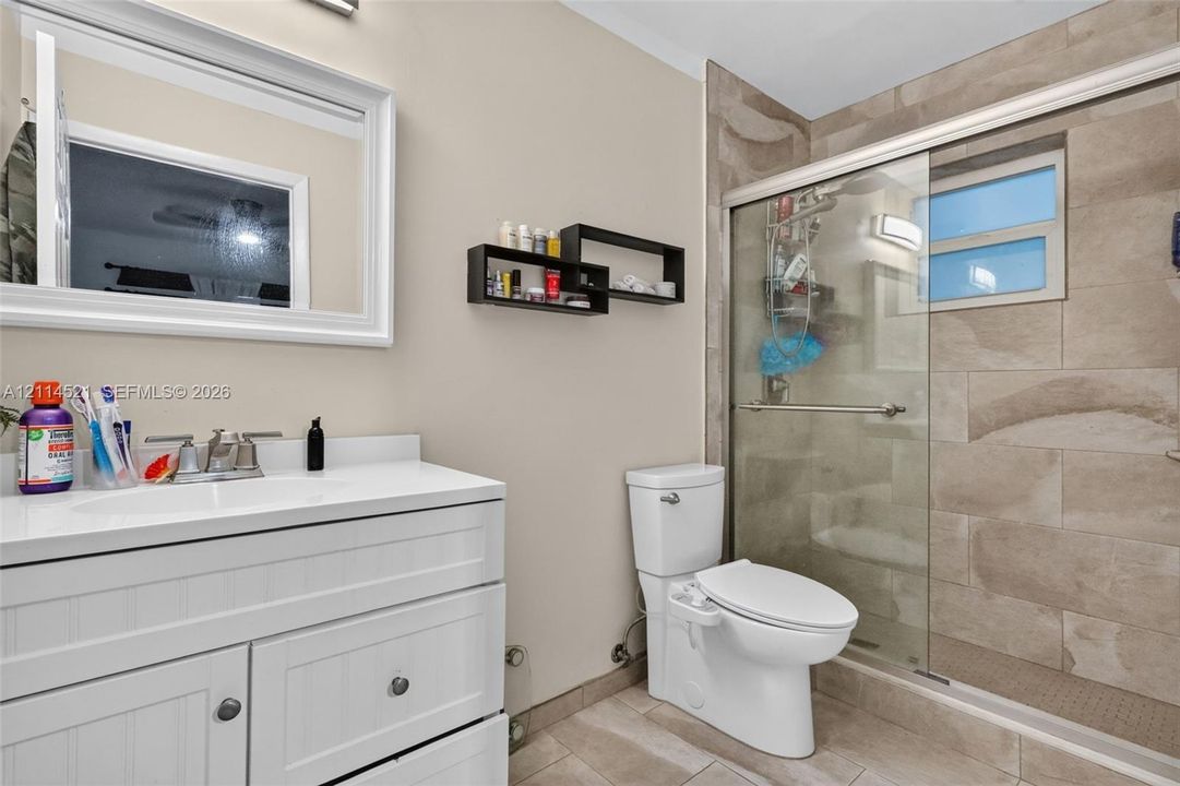 Bathroom featuring a white shaker-style vanity with an integrated countertop, a framed rectangular mirror, and a walk-in shower with stone-finish tile surround, sliding glass door, and recessed lighting