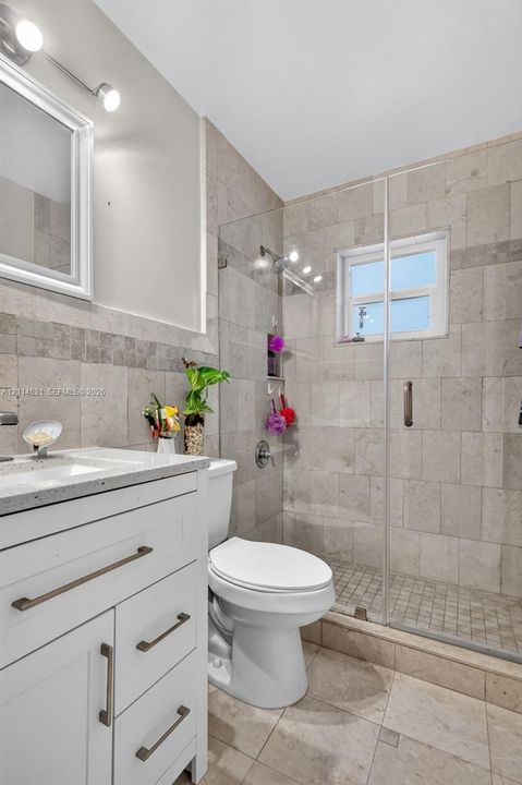 White shaker-style vanity with stone-finish countertop, frameless glass shower enclosure, full-height wall tiling, and dual-light vanity fixture