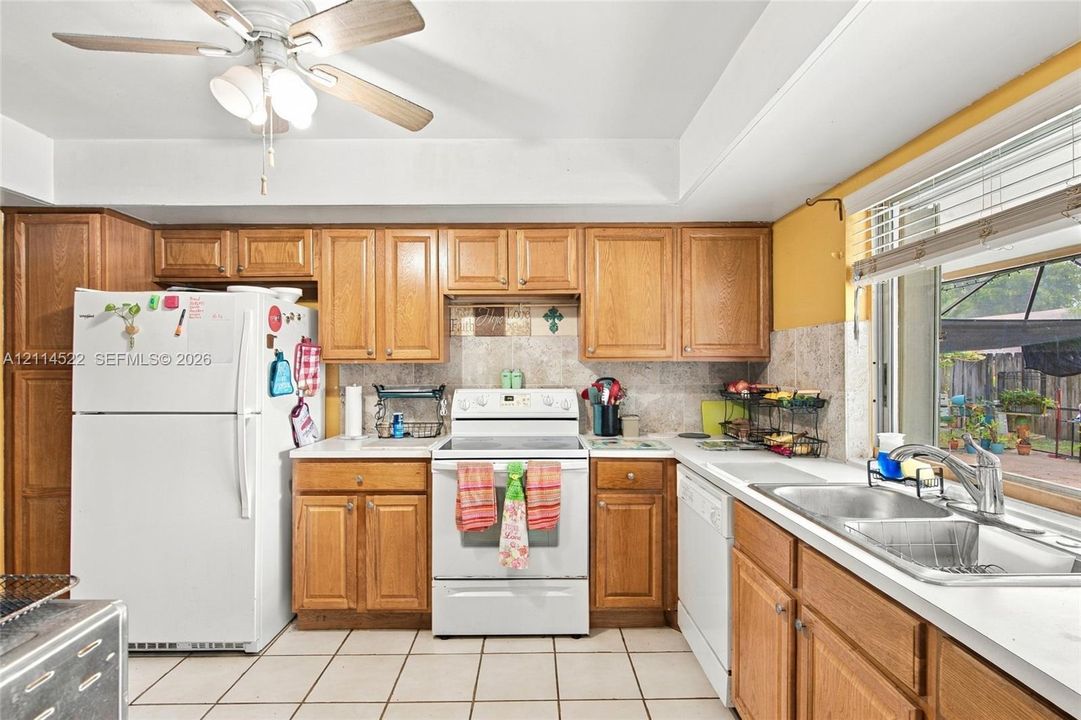 Kitchen featuring wood cabinetry, white tile flooring, tiled backsplash, stainless steel dual-basin sink, and a ceiling-mounted fan with integrated lighting