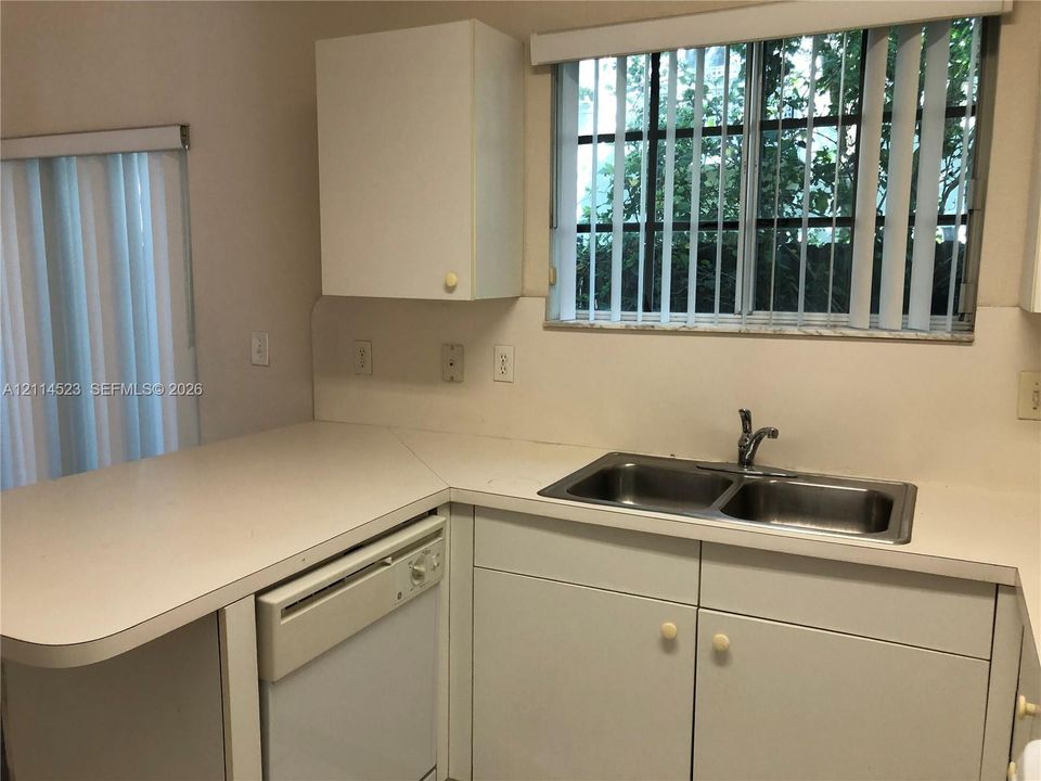 Kitchen area featuring solid-surface countertops, dual-basin stainless steel sink with metal faucet, white cabinetry, and built-in dishwasher