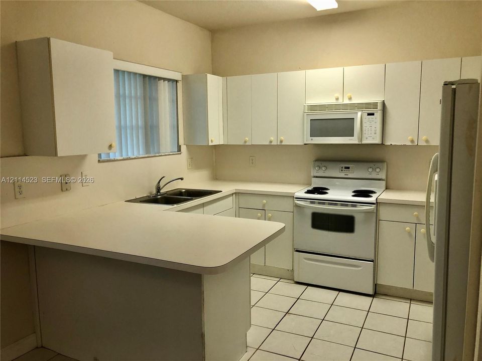 Kitchen featuring white cabinetry, solid-surface countertops, dual-basin stainless steel sink, over-the-range microwave, and ceramic tile flooring