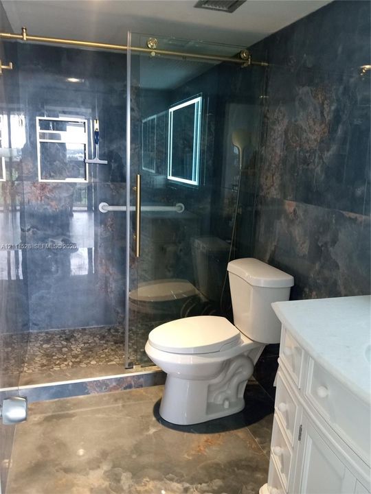 Walk-in shower featuring gold-tone hardware, a glass sliding door, and a pebble-stone floor, complemented by dark-toned tile walls, a vanity with a light-toned countertop, and a wall-mounted light-up mirror