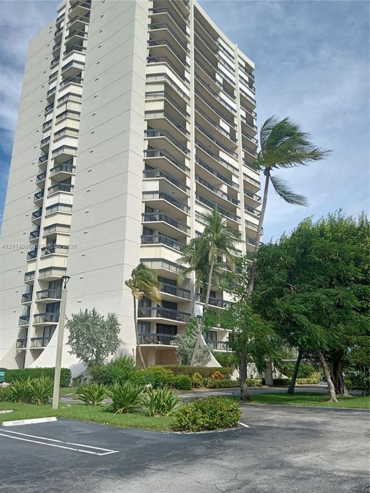 High-rise residential building featuring stacked private balconies, concrete exterior cladding, and ground-level landscaping