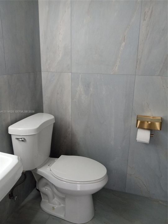 Full-height large-format wall and floor tiling with subtle veining, dual-flush toilet, and metallic gold-tone toilet paper dispenser