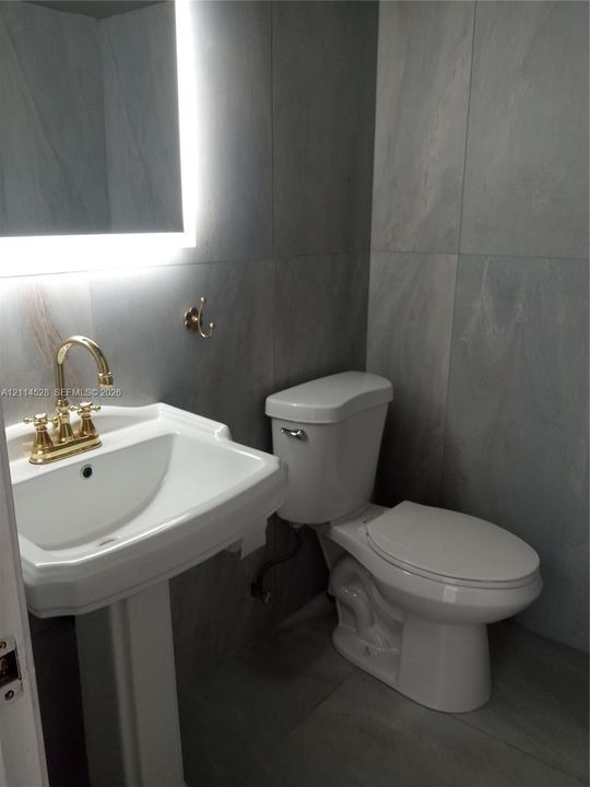 Pedestal sink with brass finish faucet, large-format grey wall tiling, and backlit vanity mirror