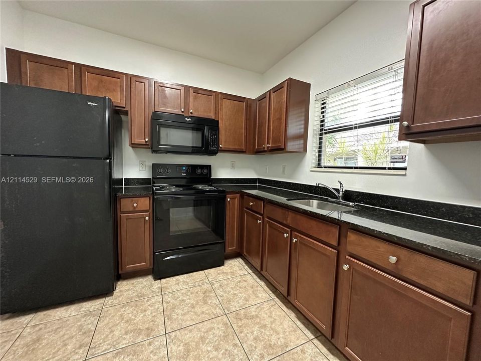 Kitchen featuring wood cabinetry, dark stone-finish countertops, black built-in appliances, ceramic tile flooring, and a stainless steel sink with a high-arch faucet
