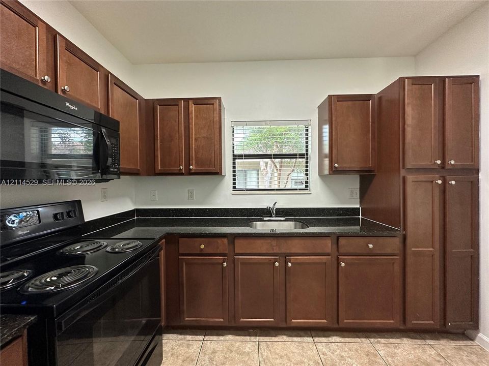 Kitchen featuring stained-wood cabinetry, dark stone-finish countertops, black built-in appliances, stainless steel undermount sink, and tile flooring