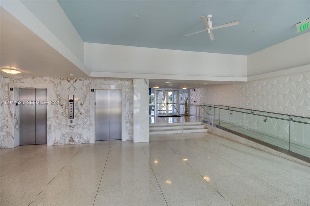 Lobby area featuring dual stainless-steel elevators, stone-paneled accent walls, polished stone flooring, a glass-panel ramp railing, and textured wall detailing