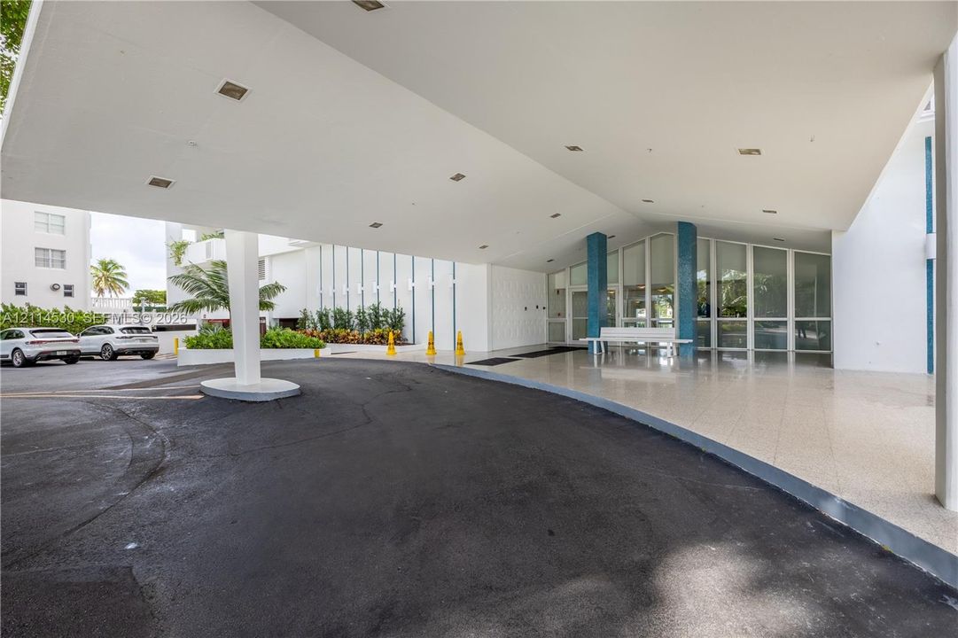 Covered porte-cochère entry featuring recessed lighting, a paved drop-off area, blue decorative columns, and full-height glass storefront glazing