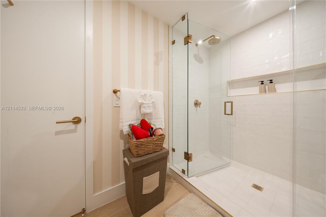 Glass-enclosed shower with white tiled walls, built-in shelving niche, brass hardware, and neutral-toned vertical striped wallpaper