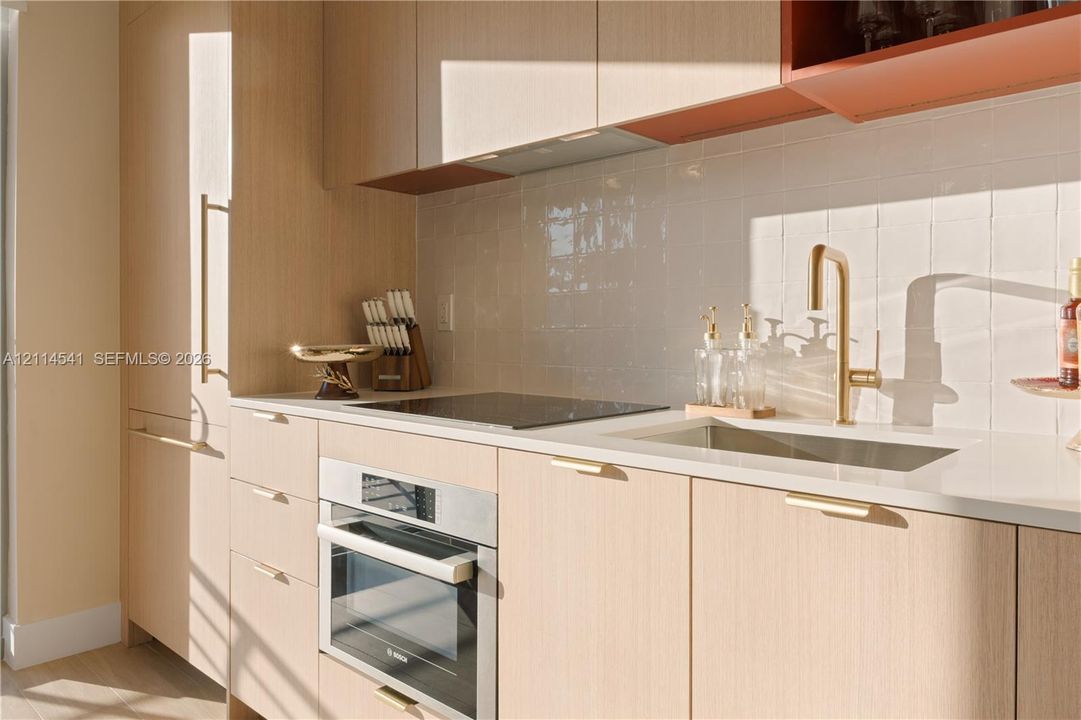 Flat-panel light-finish wood cabinetry with integrated hardware, white quartz countertops, high-gloss square tile backsplash, stainless steel built-in oven, and a brass-tone gooseneck faucet