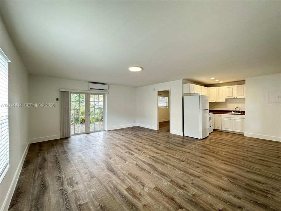 Open-concept interior featuring wood-finish flooring, glass-paned double doors, wall-mounted climate control unit, and white cabinetry with integrated kitchen appliances