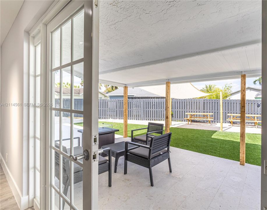 Covered patio with light-toned stone tile flooring, wooden support columns, white-framed glass doors, artificial turf, and a perimeter fence