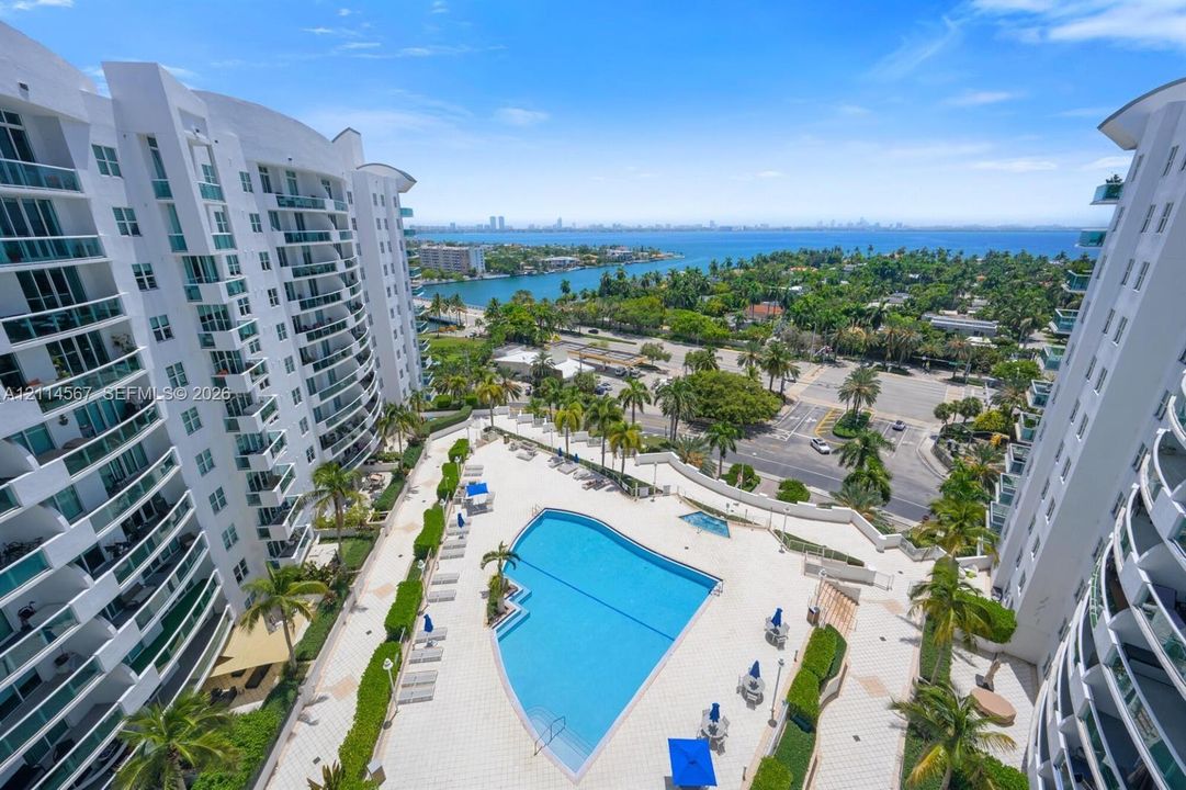 Outdoor swimming pool, paved sundeck, mature palm trees, multi-story building architecture with balconies, and coastal water backdrop