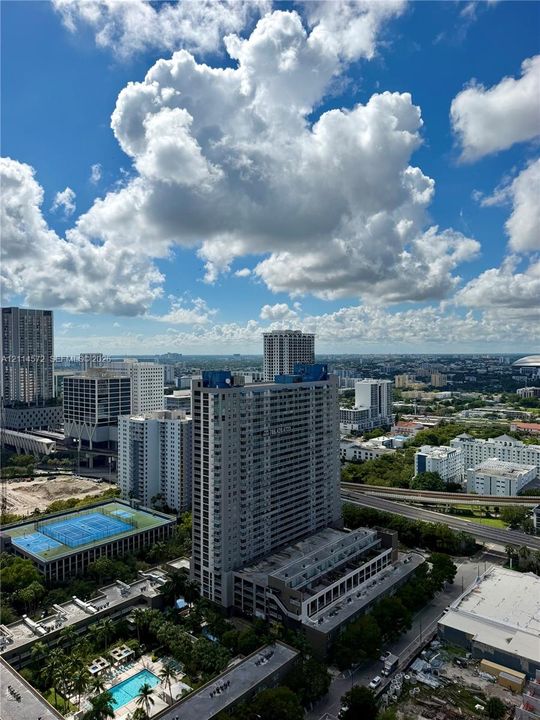 Aerial perspective of urban landscape featuring high-rise architecture, a multi-story parking structure, a blue-surface tennis court, and a rectangular ground-level pool