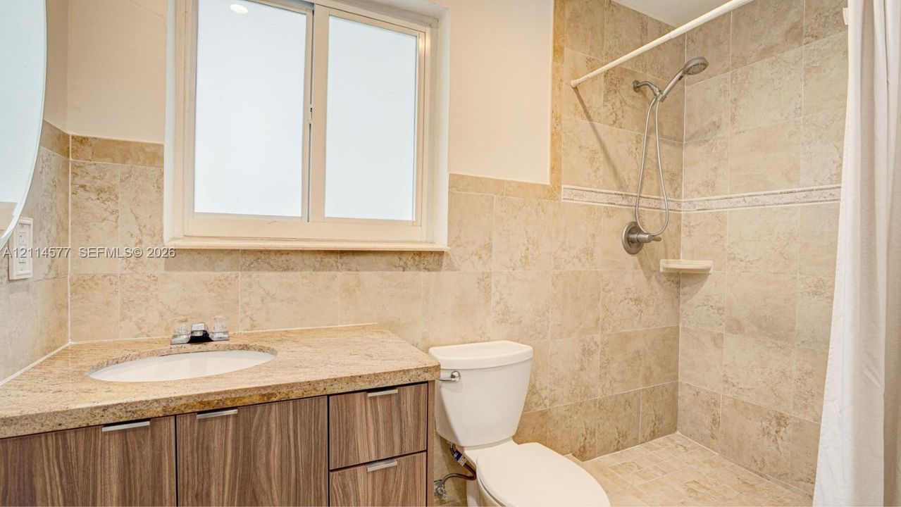 Bathroom featuring stone-tile wall surround, wood-finish vanity cabinetry, stone-slab countertop with integrated oval sink, and a handheld shower fixture