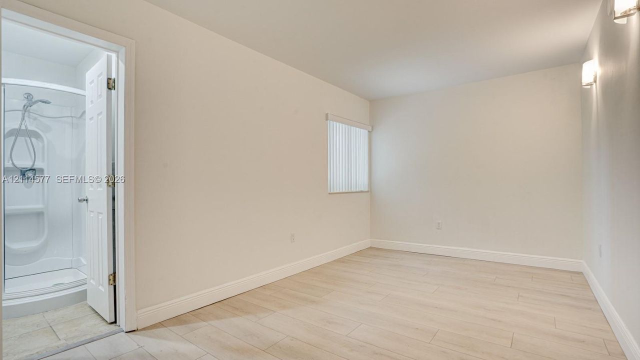 Neutral-toned room with wood-finish flooring, a vertical blind window covering, wall-mounted light fixtures, and a shower stall enclosure with a handheld faucet