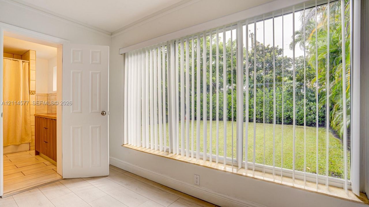 Expansive multi-pane window with vertical blinds, light-toned tile flooring, and a solid-core interior door leading to a bathroom featuring a wooden vanity and ceramic wall tiling