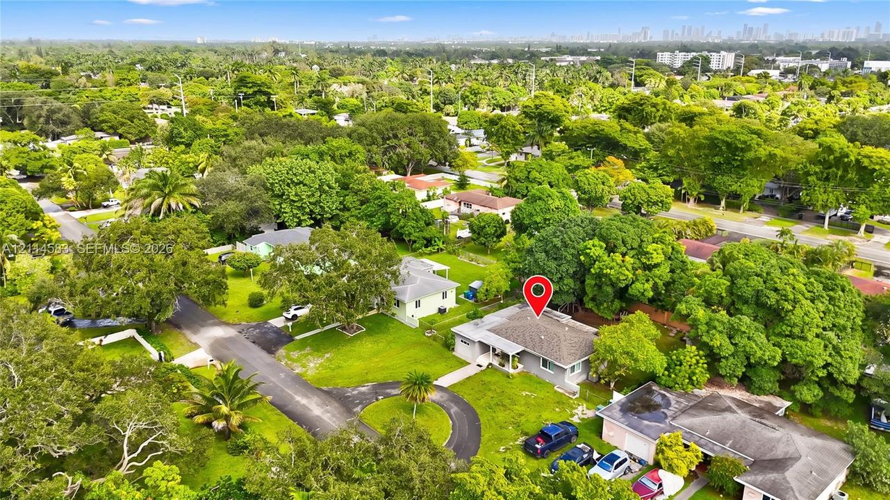Aerial perspective featuring a single-story residence with a pitched shingle roof, paved circular driveway, expansive lawn, and mature trees