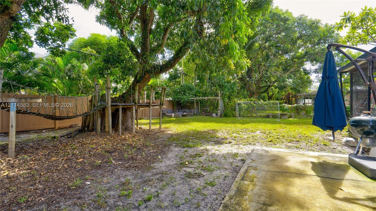 Expansive backyard featuring a concrete patio, mature trees, a wooden perimeter fence, and a wooden elevated platform structure