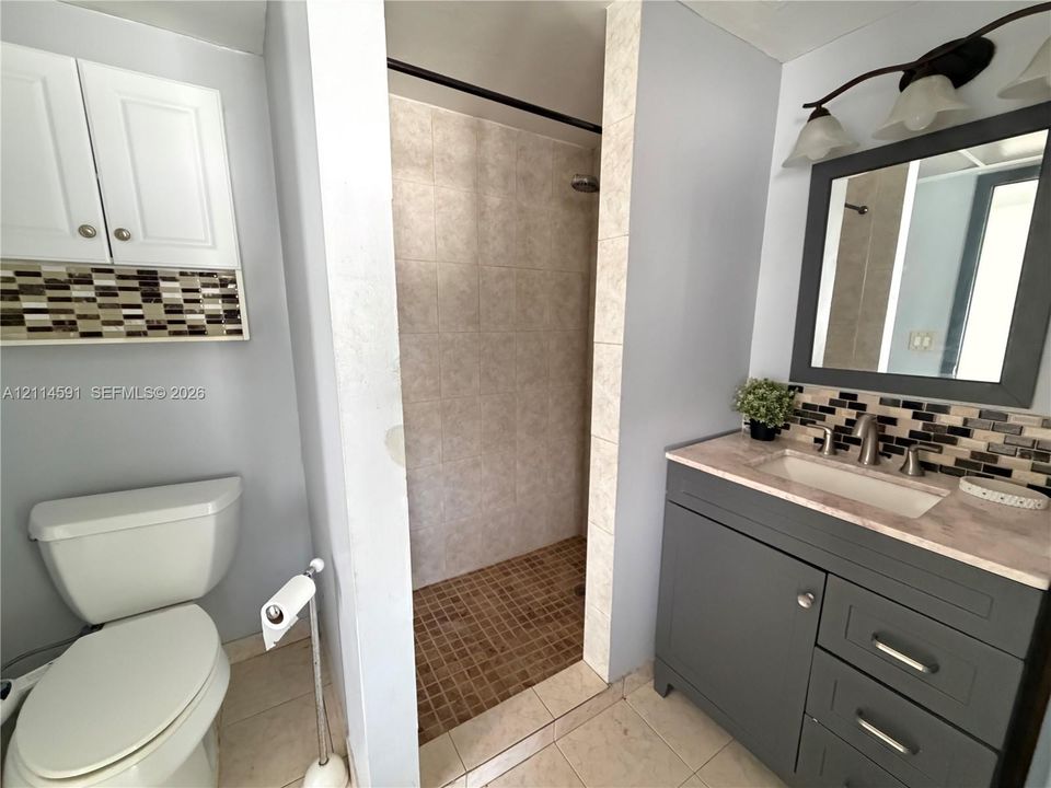 Bathroom featuring a grey vanity with stone-style countertop, integrated sink, tiled shower enclosure, multi-toned decorative backsplash, and wall-mounted vanity lighting