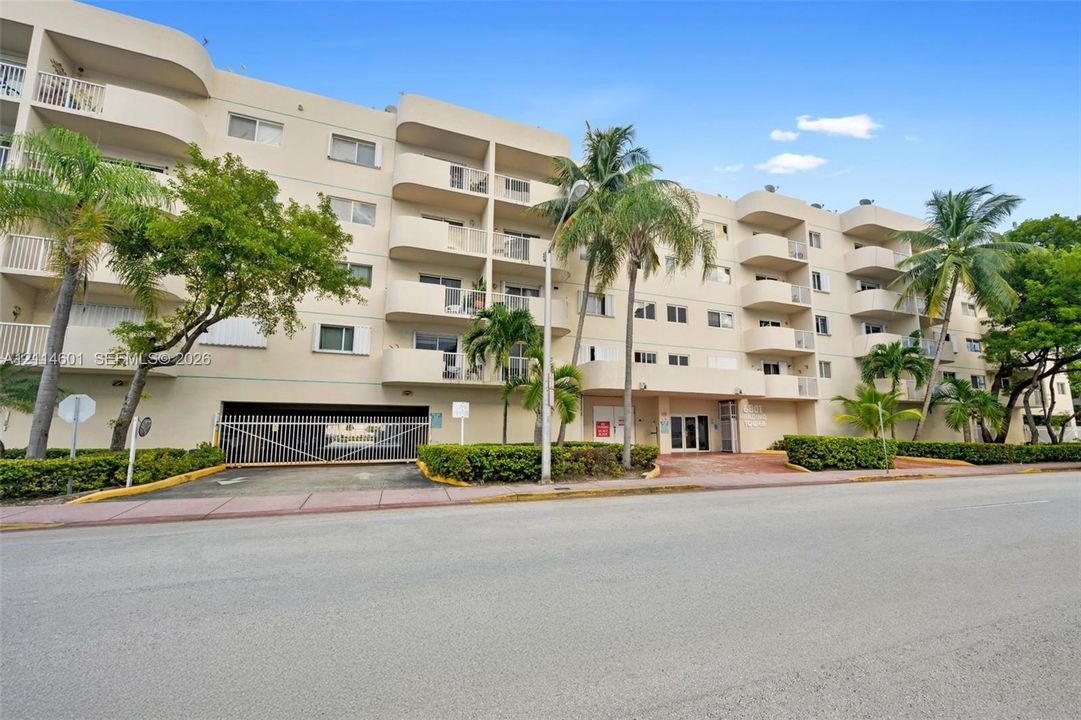 Multi-story building exterior featuring rounded balconies, secure gated parking access, and tropical landscaping