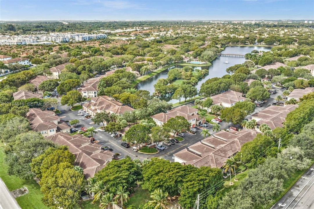 Aerial perspective of a multi-unit residential community featuring clay-tile roofing, established landscaping, paved parking areas, and a central water feature with a pedestrian bridge