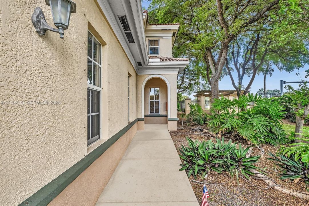 Stucco exterior with a concrete walkway, arched architectural detailing, metal wall-mounted light fixture, and mature landscape plantings