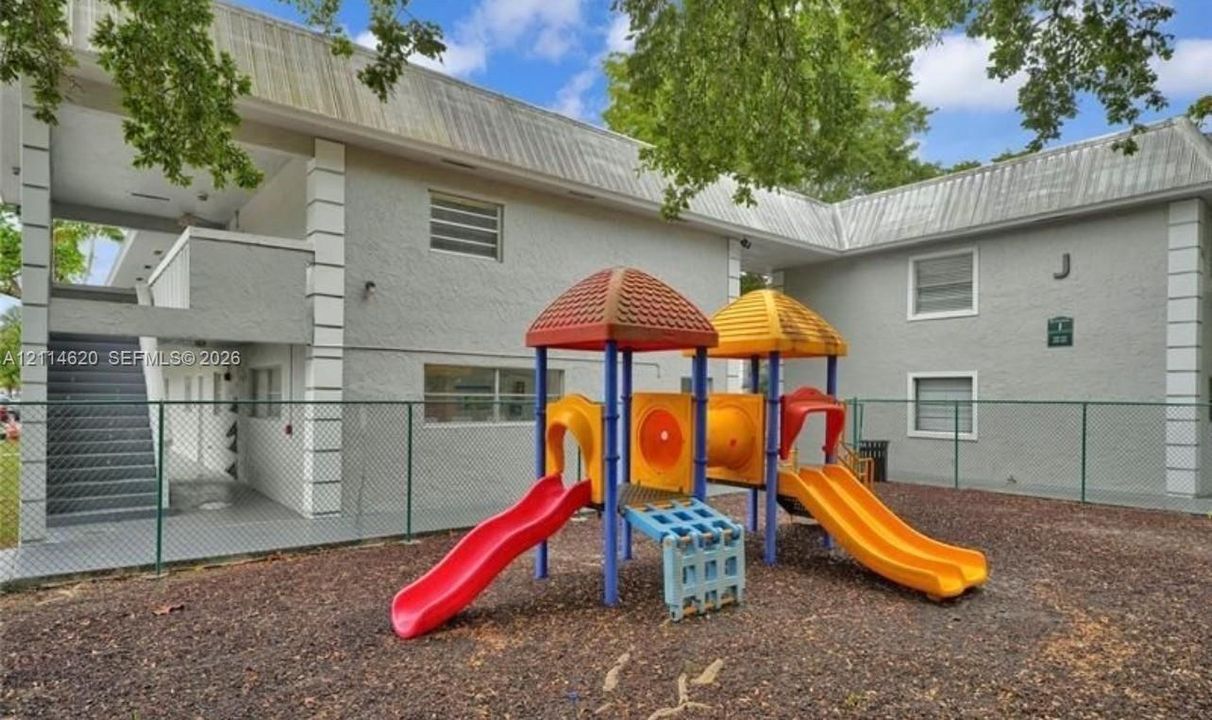Multi-story stucco exterior building featuring a fenced-in recreation area with a modular play structure and wood-chip ground cover