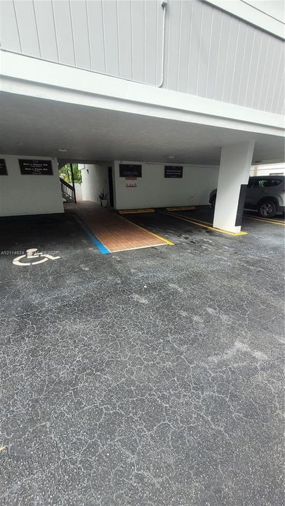 Covered parking area featuring asphalt surfacing, painted structural support columns, designated parking stalls, and a brick-paved walkway