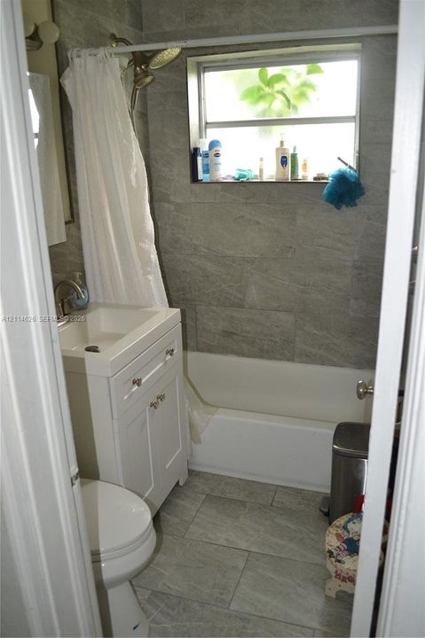 Bathroom featuring large-format gray tile flooring and wall surfacing, white vanity cabinet with a rectangular basin, and a combination bathtub and shower enclosure with a horizontal window