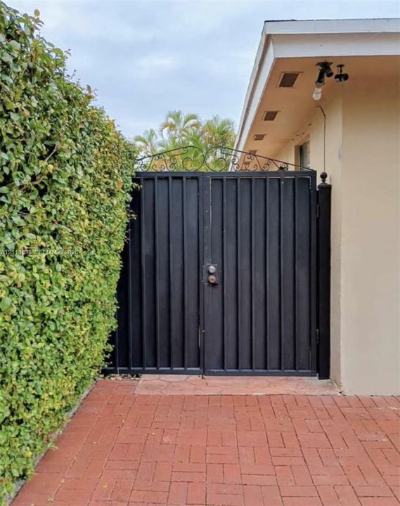 Black metal double gate featuring decorative scrollwork, installed on a brick-paved patio bordered by a dense manicured hedge