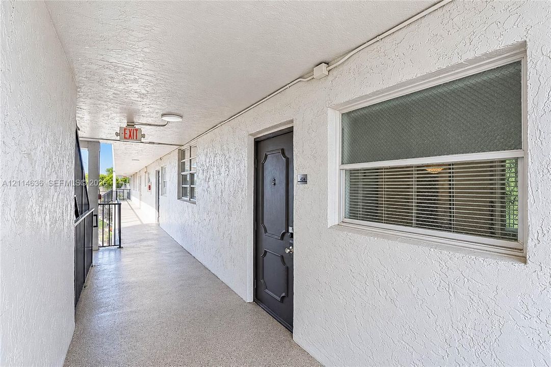 Exterior corridor featuring textured stucco walls, a paneled entry door, window with integrated blinds, and metal railing