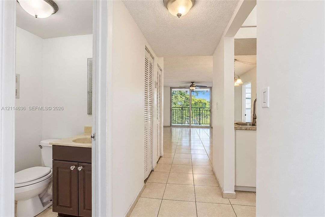 Hallway featuring neutral tile flooring, louvered-door closets, a recessed bathroom with a dark wood-finish vanity, and a glass-paneled door opening to a balcony