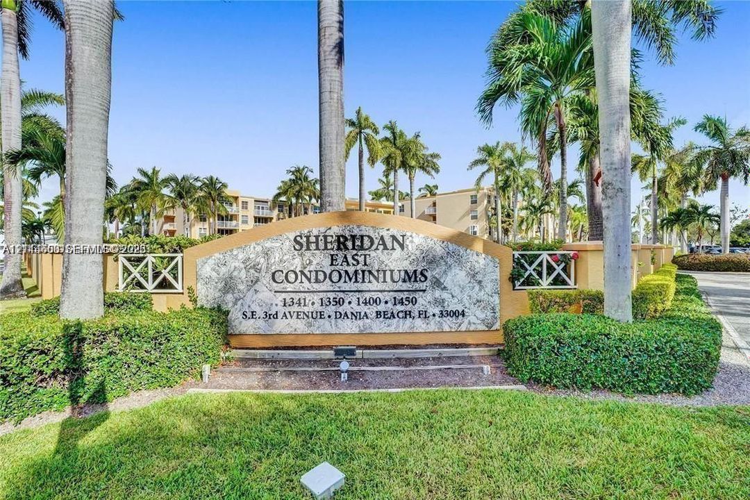 Entrance monument featuring stone-finish signage, manicured landscaping, perimeter fencing, and mature palm trees