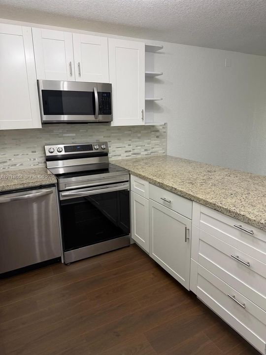 Kitchen featuring white shaker-style cabinetry, stone-finish countertops, stainless steel appliances, a multi-tone tile backsplash, and wood-finish flooring