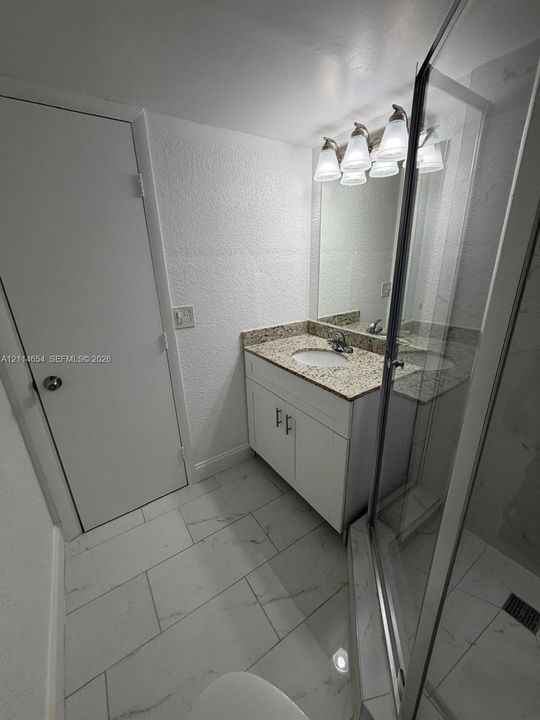 Bathroom featuring white cabinetry, stone-finish countertop with integrated oval sink, glass-enclosed shower, polished light-colored tile flooring, and multi-bulb vanity lighting