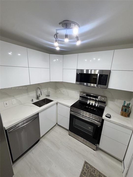 Modern kitchen featuring high-gloss white flat-panel cabinetry, light-toned stone-finish countertops, stainless steel appliances, and a contemporary multi-bulb ceiling fixture