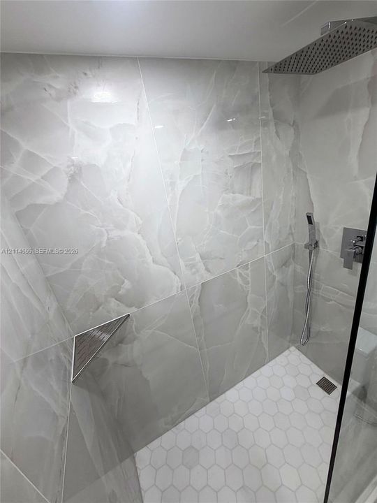 Walk-in shower featuring large-format stone-look wall tiling, white hexagonal floor tiling, a ceiling-mounted rainfall showerhead, a wall-mounted handheld sprayer, and a recessed corner shelf